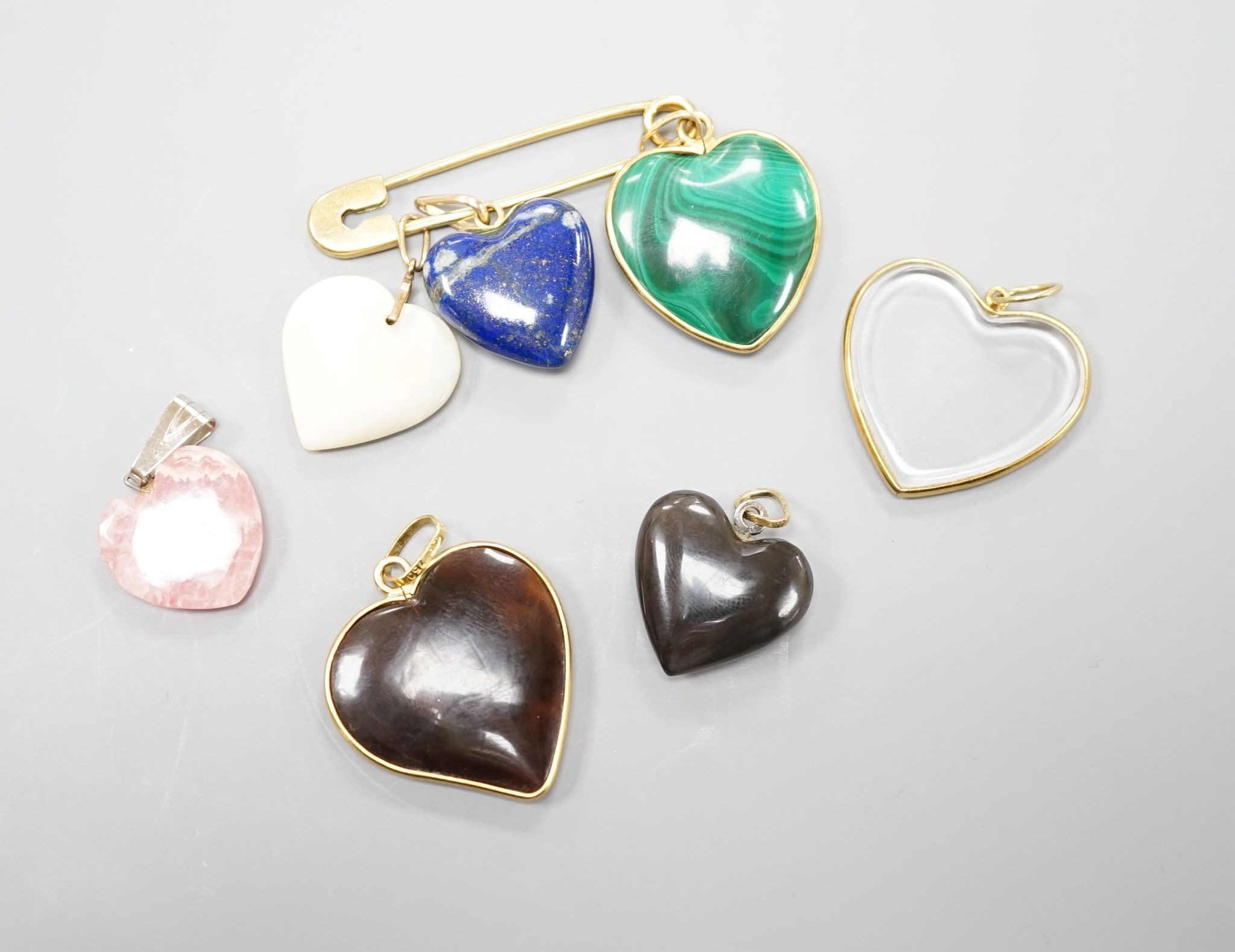 An 18ct safety pin, 47mm, hung with seven assorted heart shaped hardstone pendants or charms, two with Italian 750 yellow metal mounts.